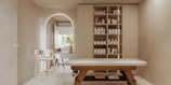A minimalist, serene spa room features a massage table with wooden legs and a white cushioned surface. To the right, a wall-mounted cabinet with open shelves holds neatly organized spa products, including bottles and jars. In the background, an archway leads to another area with a simple chair and a countertop that holds decorative items. Soft, neutral tones dominate the space, creating a tranquil atmosphere.