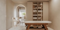A minimalist, serene spa room features a massage table with wooden legs and a white cushioned surface. To the right, a wall-mounted cabinet with open shelves holds neatly organized spa products, including bottles and jars. In the background, an archway leads to another area with a simple chair and a countertop that holds decorative items. Soft, neutral tones dominate the space, creating a tranquil atmosphere.