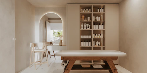 A minimalist, serene spa room features a massage table with wooden legs and a white cushioned surface. To the right, a wall-mounted cabinet with open shelves holds neatly organized spa products, including bottles and jars. In the background, an archway leads to another area with a simple chair and a countertop that holds decorative items. Soft, neutral tones dominate the space, creating a tranquil atmosphere.