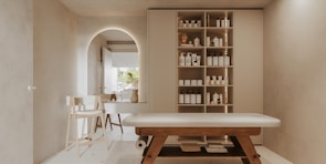 A minimalist, serene spa room features a massage table with wooden legs and a white cushioned surface. To the right, a wall-mounted cabinet with open shelves holds neatly organized spa products, including bottles and jars. In the background, an archway leads to another area with a simple chair and a countertop that holds decorative items. Soft, neutral tones dominate the space, creating a tranquil atmosphere.