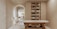 A minimalist, serene spa room features a massage table with wooden legs and a white cushioned surface. To the right, a wall-mounted cabinet with open shelves holds neatly organized spa products, including bottles and jars. In the background, an archway leads to another area with a simple chair and a countertop that holds decorative items. Soft, neutral tones dominate the space, creating a tranquil atmosphere.