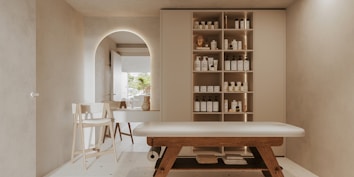 A minimalist, serene spa room features a massage table with wooden legs and a white cushioned surface. To the right, a wall-mounted cabinet with open shelves holds neatly organized spa products, including bottles and jars. In the background, an archway leads to another area with a simple chair and a countertop that holds decorative items. Soft, neutral tones dominate the space, creating a tranquil atmosphere.