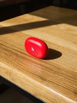 A red, oval-shaped case with a white lowercase 'b' logo on it sits centered on a wooden surface, illuminated by natural sunlight casting a shadow.