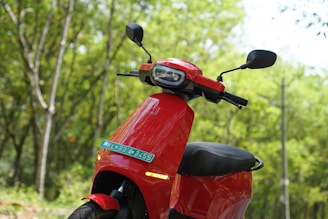 A bright red scooter with a modern design, parked in a lush green forest. The scooter's mirrors and handlebars are visible, and there is a license plate attached at the front. The background is a blur of vibrant green leaves and tree trunks.