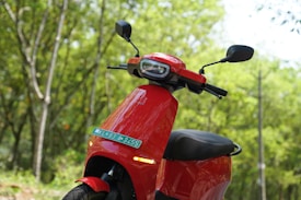 A bright red scooter with a modern design, parked in a lush green forest. The scooter's mirrors and handlebars are visible, and there is a license plate attached at the front. The background is a blur of vibrant green leaves and tree trunks.