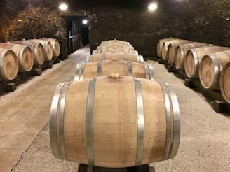 A dimly lit cellar with rows of wooden barrels lined neatly on both sides. The barrels are set on racks over a gravel floor. The walls and ceiling are made of stone, giving the atmosphere an old-world charm.