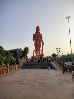 A massive statue of a figure resembling the Hindu deity Hanuman stands prominently on a raised platform with a staircase leading up to it. The statue is reddish-orange and towers over visitors who are gathered at its base. Surrounding the platform are trees and lamp posts, and the sky is clear.
