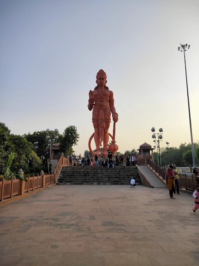 A massive statue of a figure resembling the Hindu deity Hanuman stands prominently on a raised platform with a staircase leading up to it. The statue is reddish-orange and towers over visitors who are gathered at its base. Surrounding the platform are trees and lamp posts, and the sky is clear.