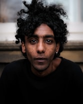 A person with curly hair and an intense gaze looks directly at the camera. The background is blurred, featuring soft neutral tones. Their expression is serious and contemplative.