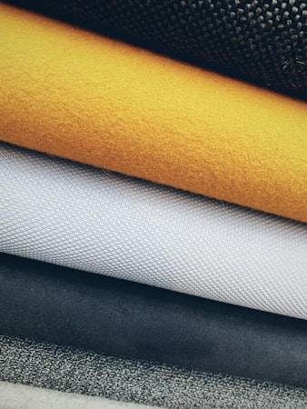 Various textured fabrics are layered on top of each other, showcasing different materials such as wool, woven textiles, and felt. The fabrics are arranged in an overlapping manner, highlighting their unique textures and patterns.