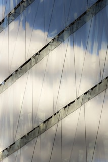 Reflections of clouds are seen on the surface of a glass building, intersected by diagonal lines and metal framework.
