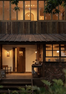 A rustic two-story wooden building with warm glowing lights visible through the windows. The lower level has a wooden entrance with a small porch area, featuring a sign and a chair. Decorative plants surround the porch, adding to the cozy ambiance.