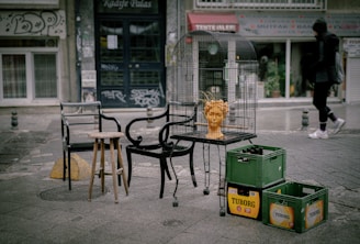 An outdoor urban scene featuring a collection of mismatched chairs and a small stool surrounding a birdcage. Inside the cage, there is an orange bust of a head with wire-like extensions protruding from it. Nearby, plastic crates with 'TUBORG' branding are stacked. The background includes a street with visible shops and one pedestrian walking by. Graffiti and urban signage are evident on the buildings.