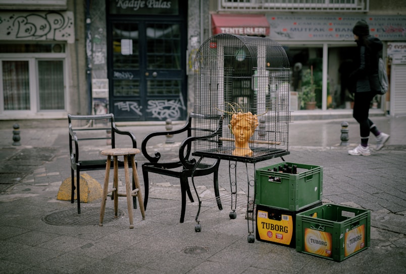 An outdoor urban scene featuring a collection of mismatched chairs and a small stool surrounding a birdcage. Inside the cage, there is an orange bust of a head with wire-like extensions protruding from it. Nearby, plastic crates with 'TUBORG' branding are stacked. The background includes a street with visible shops and one pedestrian walking by. Graffiti and urban signage are evident on the buildings.