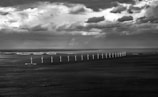 A row of wind turbines stretches across the sea under a dramatic sky filled with dark, patchy clouds. In the distance, a coastline with large buildings is visible, contrasting with the vast expanse of water and sky.