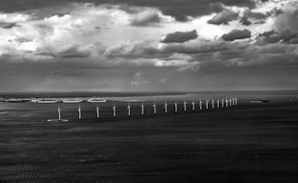 A row of wind turbines stretches across the sea under a dramatic sky filled with dark, patchy clouds. In the distance, a coastline with large buildings is visible, contrasting with the vast expanse of water and sky.
