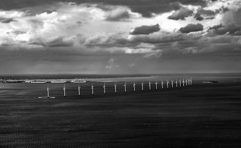 A row of wind turbines stretches across the sea under a dramatic sky filled with dark, patchy clouds. In the distance, a coastline with large buildings is visible, contrasting with the vast expanse of water and sky.