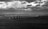A row of wind turbines stretches across the sea under a dramatic sky filled with dark, patchy clouds. In the distance, a coastline with large buildings is visible, contrasting with the vast expanse of water and sky.