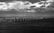 A row of wind turbines stretches across the sea under a dramatic sky filled with dark, patchy clouds. In the distance, a coastline with large buildings is visible, contrasting with the vast expanse of water and sky.