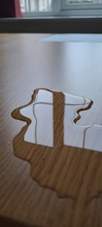 A puddle of water lies on a wooden surface, reflecting a window with multiple panes. The focus is on the water and the reflection, while the background includes a blurred view of a window frame.