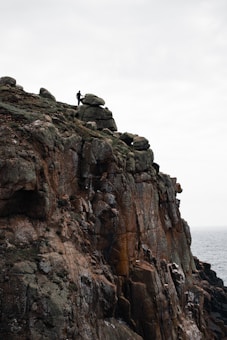 A lone figure stands atop a rugged cliff covered in large, jagged rocks, overlooking the sea. The overcast sky adds to the dramatic ambiance of the scene.