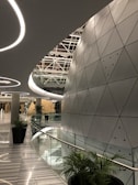 A modern architectural space with futuristic design elements, featuring a large, curved wall with triangular metallic panels. The ceiling has circular light fixtures and a geometric pattern. Tall green plants are placed along the glass railing that borders a curved corridor, leading to a round, dome-like structure in the distance.
