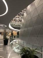 A modern architectural space with futuristic design elements, featuring a large, curved wall with triangular metallic panels. The ceiling has circular light fixtures and a geometric pattern. Tall green plants are placed along the glass railing that borders a curved corridor, leading to a round, dome-like structure in the distance.