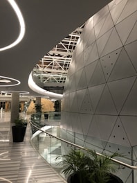 A modern architectural space with futuristic design elements, featuring a large, curved wall with triangular metallic panels. The ceiling has circular light fixtures and a geometric pattern. Tall green plants are placed along the glass railing that borders a curved corridor, leading to a round, dome-like structure in the distance.