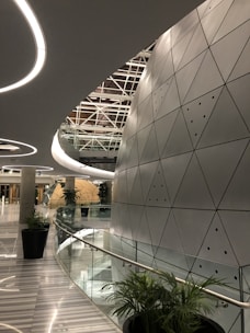 A modern architectural space with futuristic design elements, featuring a large, curved wall with triangular metallic panels. The ceiling has circular light fixtures and a geometric pattern. Tall green plants are placed along the glass railing that borders a curved corridor, leading to a round, dome-like structure in the distance.
