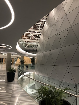 A modern architectural space with futuristic design elements, featuring a large, curved wall with triangular metallic panels. The ceiling has circular light fixtures and a geometric pattern. Tall green plants are placed along the glass railing that borders a curved corridor, leading to a round, dome-like structure in the distance.