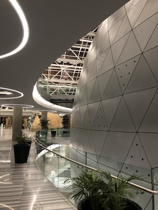A modern architectural space with futuristic design elements, featuring a large, curved wall with triangular metallic panels. The ceiling has circular light fixtures and a geometric pattern. Tall green plants are placed along the glass railing that borders a curved corridor, leading to a round, dome-like structure in the distance.