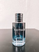 A bottle of Dior Sauvage cologne is placed on a dark surface. The bottle is transparent, showcasing the light blue liquid inside. The cap is metallic and cylindrical with a sleek design. Reflective elements are visible on the bottle's surface.