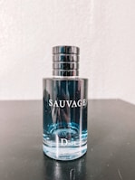 A bottle of Dior Sauvage cologne is placed on a dark surface. The bottle is transparent, showcasing the light blue liquid inside. The cap is metallic and cylindrical with a sleek design. Reflective elements are visible on the bottle's surface.