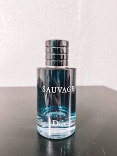 A bottle of Dior Sauvage cologne is placed on a dark surface. The bottle is transparent, showcasing the light blue liquid inside. The cap is metallic and cylindrical with a sleek design. Reflective elements are visible on the bottle's surface.