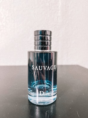 A bottle of Dior Sauvage cologne is placed on a dark surface. The bottle is transparent, showcasing the light blue liquid inside. The cap is metallic and cylindrical with a sleek design. Reflective elements are visible on the bottle's surface.
