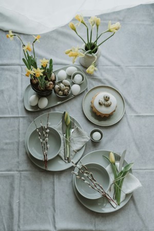 A table set for a spring-themed gathering with white and speckled eggs arranged in a light green platter, complemented by vases filled with yellow daffodils. Plates are neatly set with silverware and adorned with budding branches and yellow tulips. A small cake with icing and three speckled eggs on top is placed beside the floral arrangements. Soft lighting enhances the pastel and earthy tones, creating an elegant yet cozy atmosphere.