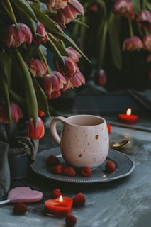 A cozy and intimate setting featuring a white mug with a hole pattern sitting on a gray plate, surrounded by fresh raspberries. Bright pink tulips drape over, adding a touch of natural beauty. Heart-shaped candles and a lollipop enhance the romantic ambiance.