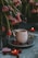 A cozy and intimate setting featuring a white mug with a hole pattern sitting on a gray plate, surrounded by fresh raspberries. Bright pink tulips drape over, adding a touch of natural beauty. Heart-shaped candles and a lollipop enhance the romantic ambiance.