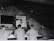 A group of men in military uniforms are gathered in a rustic attic-like room, where they seem to be engaged in a lesson or lecture. One man stands at the front, pointing at an anatomical chart displayed on the wall, while the others are seated at benches, attentively observing.