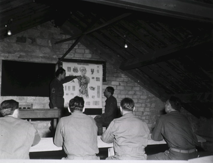 A group of men in military uniforms are gathered in a rustic attic-like room, where they seem to be engaged in a lesson or lecture. One man stands at the front, pointing at an anatomical chart displayed on the wall, while the others are seated at benches, attentively observing.