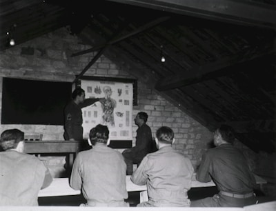 A group of men in military uniforms are gathered in a rustic attic-like room, where they seem to be engaged in a lesson or lecture. One man stands at the front, pointing at an anatomical chart displayed on the wall, while the others are seated at benches, attentively observing.