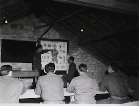 A group of men in military uniforms are gathered in a rustic attic-like room, where they seem to be engaged in a lesson or lecture. One man stands at the front, pointing at an anatomical chart displayed on the wall, while the others are seated at benches, attentively observing.