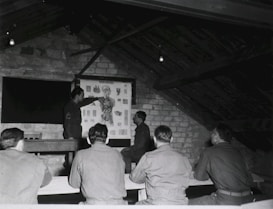 A group of men in military uniforms are gathered in a rustic attic-like room, where they seem to be engaged in a lesson or lecture. One man stands at the front, pointing at an anatomical chart displayed on the wall, while the others are seated at benches, attentively observing.