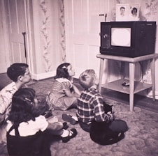 Four children sit on the carpet, engrossed in watching a black and white television placed on a wooden stand. The room has floral wallpaper and a carpeted floor. The children, appearing to be from the mid-20th century based on their clothing, display attentive postures, showcasing a sense of curiosity and engagement.