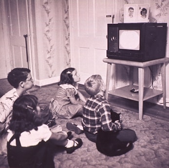 Four children sit on the carpet, engrossed in watching a black and white television placed on a wooden stand. The room has floral wallpaper and a carpeted floor. The children, appearing to be from the mid-20th century based on their clothing, display attentive postures, showcasing a sense of curiosity and engagement.