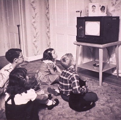 Four children sit on the carpet, engrossed in watching a black and white television placed on a wooden stand. The room has floral wallpaper and a carpeted floor. The children, appearing to be from the mid-20th century based on their clothing, display attentive postures, showcasing a sense of curiosity and engagement.