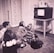 Four children sit on the carpet, engrossed in watching a black and white television placed on a wooden stand. The room has floral wallpaper and a carpeted floor. The children, appearing to be from the mid-20th century based on their clothing, display attentive postures, showcasing a sense of curiosity and engagement.