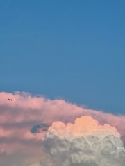 A sky scene featuring a vibrant blue background with large fluffy clouds. The clouds are illuminated with shades of pink and orange, suggesting a sunrise or sunset. A single bird is seen flying to the left of the clouds, adding a sense of scale and movement.
