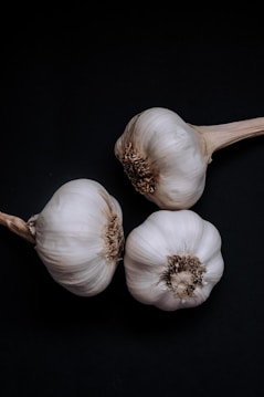 Three bulbs of garlic are arranged against a dark black background, highlighting their natural white and light brown hues. The rough texture and papery skin of the garlic are visible.