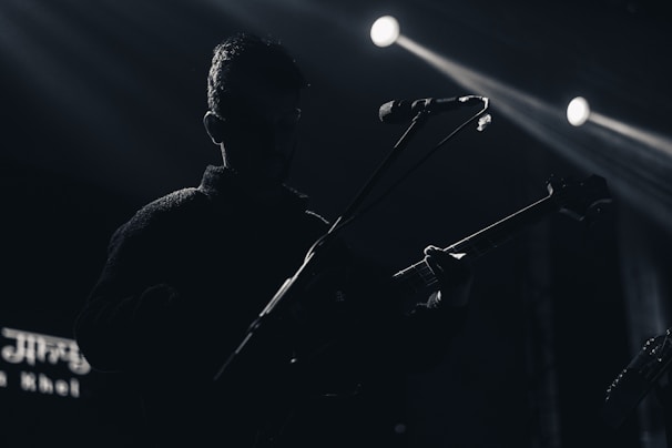 A silhouette of a musician holding an electric guitar, standing under spotlight illumination. The background is dark, with visible beams of light and a microphone positioned near the musician's face.